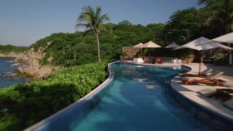 Luftaufnahme-Eines-Luxuriösen-Blauen-Swimmingpools-In-Einem-Hotel-In-Huatulco,-Umgeben-Von-Sich-Wiegenden-Palmen,-Die-Sich-Rückwärts-Bewegen