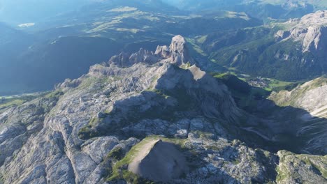 Aerial-view-of-the-Dolomites,-prominently-featuring-the-rugged-Sassongher-peak,-with-the-town-of-Kolfuschg-nestled-in-the-valley-below-and-vast-mountain-ranges-stretching-into-the-distance