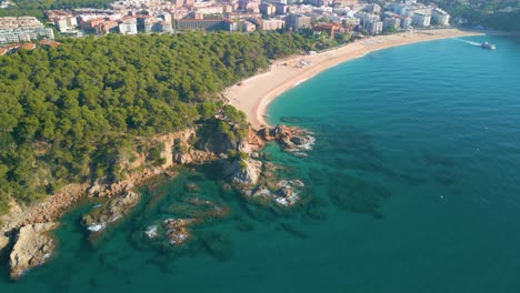 aerial-images-of-Lloret-De-Mar,-Fenals-beach-over-the-botanical-garden,-few-people-on-the-beach,-transparent-waters,-rocks-and-green-vegetation-blue-sky