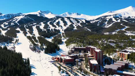 Aerial-Drone-View-of-Giant-Ski-Resort-with-People-Skiing-by-Full-Parking-Lot-and-Riding-Chairlifts-Down-Mountain-Slopes-through-a-Pine-Tree-Forest-Covered-in-Snow