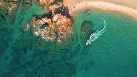 Aerial-view-of-a-beach,-with-rocks-with-calm,-super-transparent-waters,-flying-over-a-small-white-motor-boat-in-Lloret-De-Mar