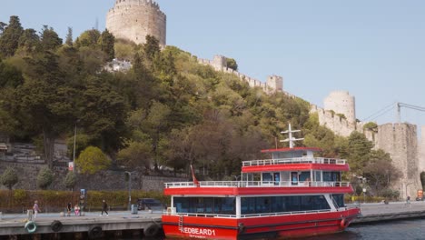 Red-tourist-cruise-boat-moored-shores-of-Bosphorus-Rumeli-Fortress