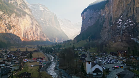 Drone-ascends-above-calm-town-in-Lauterbrunnen,-Switzerland-in-the-shadow-of-canyon-walls
