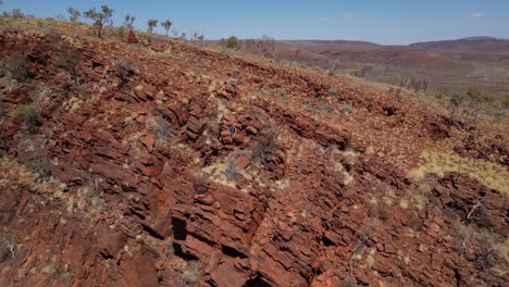 Orbiting-aerial-shot-of-man-climbing-on-red-desert-mountain-wall-during-sunny-day-in-Australia---blue-sky-in-summer-with-hiking-adventure