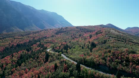 Luftaufnahmen-über-Einem-Waldgebiet-Mit-Leuchtenden-Herbstfarben,-In-Der-Nähe-Des-Kyhv-Gipfels-In-Utah,-Mit-Einer-Kurvenreichen-Straße-Und-Einer-Beeindruckenden-Bergkette-An-Einem-Klaren-Tag
