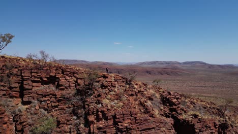 Walking-man-on-the-edge-of-red-mountains-during-sunny-day-in-Australia-desert---Aerial-view
