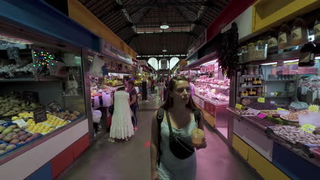 Dolly-backwards-shot-of-a-young-pretty-woman-with-a-drink-in-hand-while-walking-through-the-covered-street-market-of-malaga-in-spain-during-a-vacation-shopping-trip-in-slow-motion
