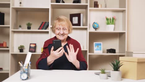 Eine-ältere-Frau-Lächelt-Freudig,-Während-Sie-Per-Videoanruf-Auf-Ihrem-Smartphone-Spricht,-Während-Sie-Am-Tisch-Sitzt