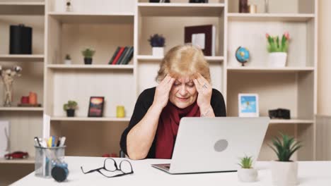 Eine-ältere,-Traurige-Frau-Sitzt-Hinter-Einem-Laptop-Und-Versucht,-Migräne-Und-Kopfschmerzen-Durch-Eine-Massage-Ihrer-Schläfen-Loszuwerden