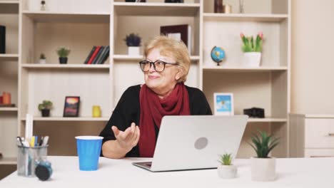 Eine-ältere-Frau-Mit-Brille-Spricht-Per-Videoanruf,-Während-Sie-Mit-Einem-Laptop-An-Einem-Tisch-Sitzt