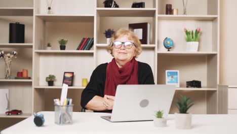 The-older-woman-in-glasses-puts-on-glasses-with-a-smile,-looking-into-the-camera-while-sitting-at-a-table-with-a-laptop