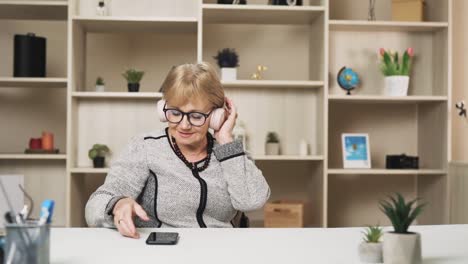 Ältere-Frau-Setzt-Kopfhörer-Auf-Und-Spielt-Musik-Auf-Ihrem-Smartphone,-Beginnt-Fröhlich-Zu-Summen-Und-Nickt-Mit-Dem-Kopf
