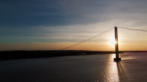 Soaring-high-above,-a-drone's-eye-view-captures-the-Humber-Bridge-bathed-in-the-warmth-of-a-setting-sun,-while-cars-gracefully-traverse-its-span,-painting-a-picture-of-tranquility-in-motion