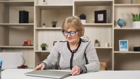 Eine-ältere-Frau-Nimmt-Ihre-Brille-Ab,-Beendet-Ihre-Arbeit-Am-Laptop-Und-Blickt-Lächelnd-In-Die-Kamera