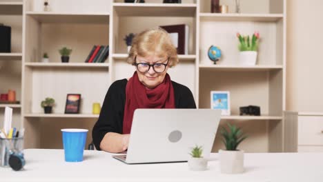 The-older-woman-in-glasses-is-working-and-drinking-a-beverage-from-a-glass-while-sitting-at-a-table-with-a-laptop