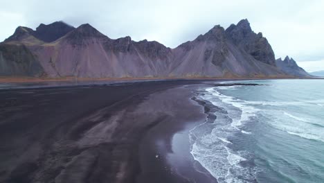 Aerial:-Vestrahorn,-with-its-iconic-jagged-peaks-and-towering-buttresses,-rises-dramatically-from-this-dark-expanse-like-a-colossal-cathedral-of-stone