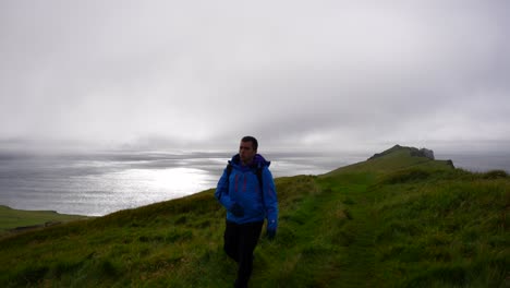 Man-walking-towards-camera-on-winter-blue-jacket-hiking-Mykines,-Faroe-Islands