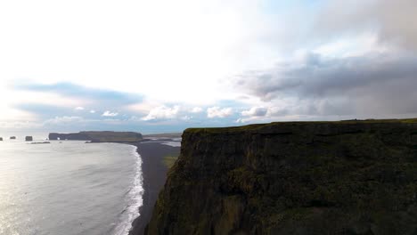 Aerial-revealing-shot-of-a-volcanic-black-sand-beach-from-behind-a-cliff-in-Iceland