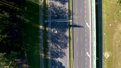 A-view-of-a-busy-highway-near-Wielki-Kack,-Gdynia,-Poland,-with-cars-rushing-past,-and-woods-casting-shadows-on-the-left-side,-seen-from-an-aerial-perspective-in-bright-sunlight