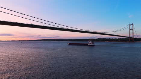 An-unhurried-barge-glides-beneath-Humber-Bridge-at-sunset,-captured-in-a-mesmerizing-aerial-drone-clip-during-the-golden-hour