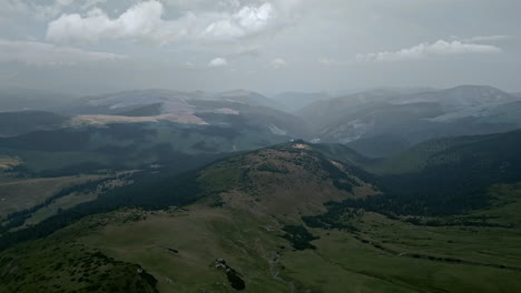 Breathtaking-aerial-view-of-Romania's-Transalpina-region,-showcasing-layered-mountains,-deep-forests,-and-open-meadows-under-a-cloud-dotted-sky