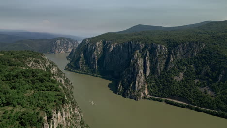 Breathtaking-view-of-Dubova,-Romania,-capturing-the-serene-river-flanked-by-majestic-limestone-cliffs,-with-a-solitary-sailboat-gliding-gracefully
