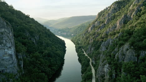 Vista-Panorámica-Del-Río-Danubio,-Flanqueada-Por-Exuberantes-Montañas-Verdes-Con-La-Icónica-Escultura-Rupestre-De-Decebalus-En-Dubova,-Rumania