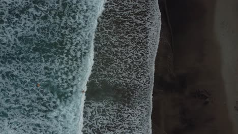 Aerial-drone-video-looking-down-on-volcanic-black-beach-and-waves-in-deep-blue-sea