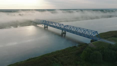 Aerial-establishing-view-of-the-steel-bridge-over-Lielupe-river-on-a-sunny-summer-morning,-fog-rising-over-the-river,-cars-driving,-wide-drone-shot-moving-forward,-tilt-down