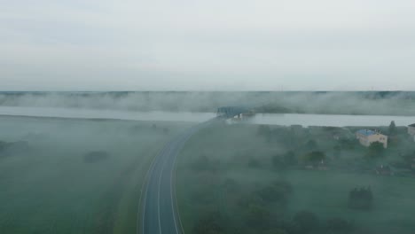 Aerial-establishing-view-of-the-steel-bridge-over-Lielupe-river-on-a-sunny-summer-morning,-fog-rising-over-the-river,-cars-driving,-wide-drone-shot-moving-forward-low