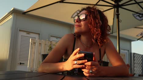 redhead-girl-in-sunglasses-enjoys-the-sunset-holding-a-smartphone-in-her-hands