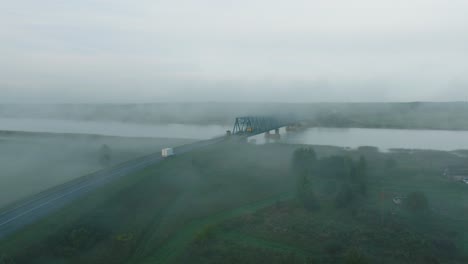 Aerial-establishing-view-of-the-steel-bridge-over-Lielupe-river-on-a-sunny-summer-morning,-fog-rising-over-the-river,-cars-driving,-wide-drone-shot-moving-forward-low