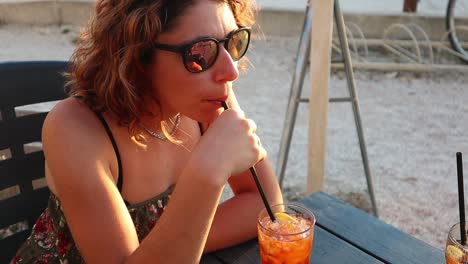 Caucasian-girl-in-sunglasses-enjoys-a-colorful-drink-at-the-breezy-sunset