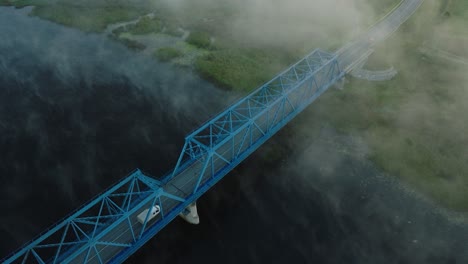Aerial-establishing-view-of-the-steel-bridge-over-Lielupe-river-on-a-sunny-summer-morning,-fog-rising-over-the-river,-cars-driving,-wide-birdseye-drone-shot-moving-forward,-tilt-down