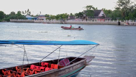 Un-Barquero-Navegando-Por-El-Río-Con-Su-Largo-Barco-De-Turismo-Mientras-Otro-Hombre-Con-Su-Barco-Es-Visto-Desde-Una-Distancia-Muy-Cerca-Del-Mercado-Flotante-De-Amphawa-En-Samut-Songkram,-Tailandia
