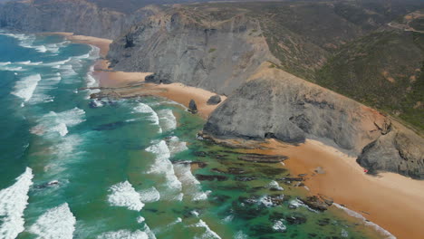 Aerial-view-of-the-paradisiacal-Vicentine-coast-with-cliffs-next-to-the-beach-and-waves-breaking-on-the-shore-on-a-sunny-summer-day,-Algarve,-Portugal