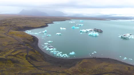 Vista-Aérea-De-Un-Lago-Glacial-Con-Hielo-Y-Un-Pintoresco-Paisaje-Montañoso-En-Islandia