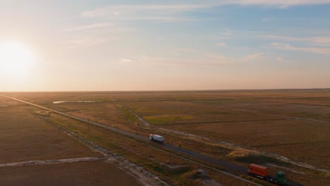Aerial-tracking-shot-of-a-transport-truck-that-navigates-a-straight-road,-forging-ahead-towards-the-radiant-embrace-of-the-setting-sun,-4K60FPS,-cinematic-grade