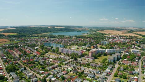 Aerial-drone-shot-of-a-czech-city-with-buldings,-cars,-roads-and-lake-with-forest-during-summer