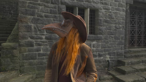 Redhead-wearing-plague-mask-and-cowboy-hat-standing-among-stone-steps-and-stone-buildings-on-bright-autumn-day