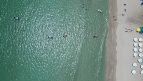 top-down-bird-view-beach-with-white-umbrellas-placed-in-line-along-the-shore-other-colourful-ones-placed-around-sea-ocean-water-waves-coming-and-going-small-vibrant-boats-anchored-close-by-holiday-up