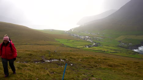 Woman-hiking-foggy-mountain-leaving-Vidareidi-village-behind,-Faroe-Islands