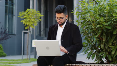 Happy-Indian-businessman-typing-working-remote-on-wireless-laptop-computer-browsing-online-outdoors