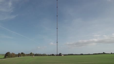 Mäusebussard-Raubvogel-Fliegt-über-Feld-In-Dänemark-Mit-Radio--Und-Fernsehturm-Im-Hintergrund---Drohnenansicht