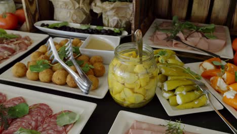 A-table-with-snacks,-on-which-there-are-plates-of-marinated-bell-peppers,-cold-cuts,-pickles,-tomatoes,-and-olives