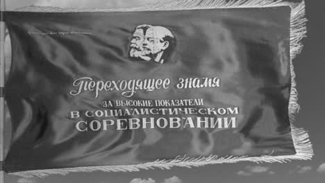 In-the-style-of-a-black-and-white-newsreel,-a-Soviet-flag-depicting-Marx-and-Lenin-flies-in-slow-motion