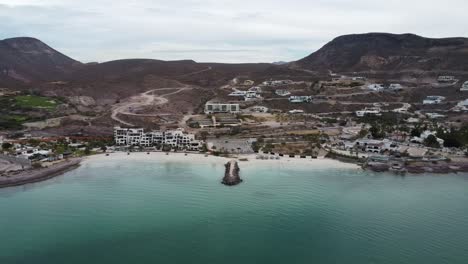 Aerial-view-of-the-wonderful-Playa-El-Caymancito-during-a-summer-trip-through-Baja-California-Sur-Mexico-with-view-of-the-beautiful-beach,-hotel-buildings-and-dry-landscape-with-mountains