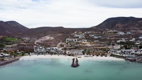 Aerial-view-in-front-of-the-dream-destination-off-the-coast-of-Playa-El-Caymancito-in-Baja-California-Sur,-Mexico-overlooking-turquoise-sea-water,-beautiful-sandy-beach-and-hotel-building-for-tourists
