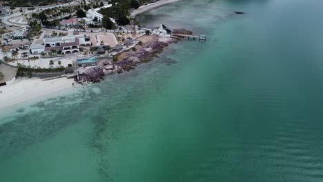 Aerial-parallax-shot-over-the-beautiful-coast-of-Playa-El-Caymancito-near-La-paz-Baja-California-Sur-Mexico-on-the-beach-with-hotel-buildings,-turquoise-sea-and-dry-summer-landscape