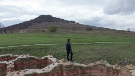 Mann-Geht-Auf-Einer-Eingestürzten-Ziegelmauer-An-Einem-Abgelegenen-Ort-In-Einer-Antiken-Stadt-In-Bulgarien,-Mit-Bergen-Im-Hintergrund